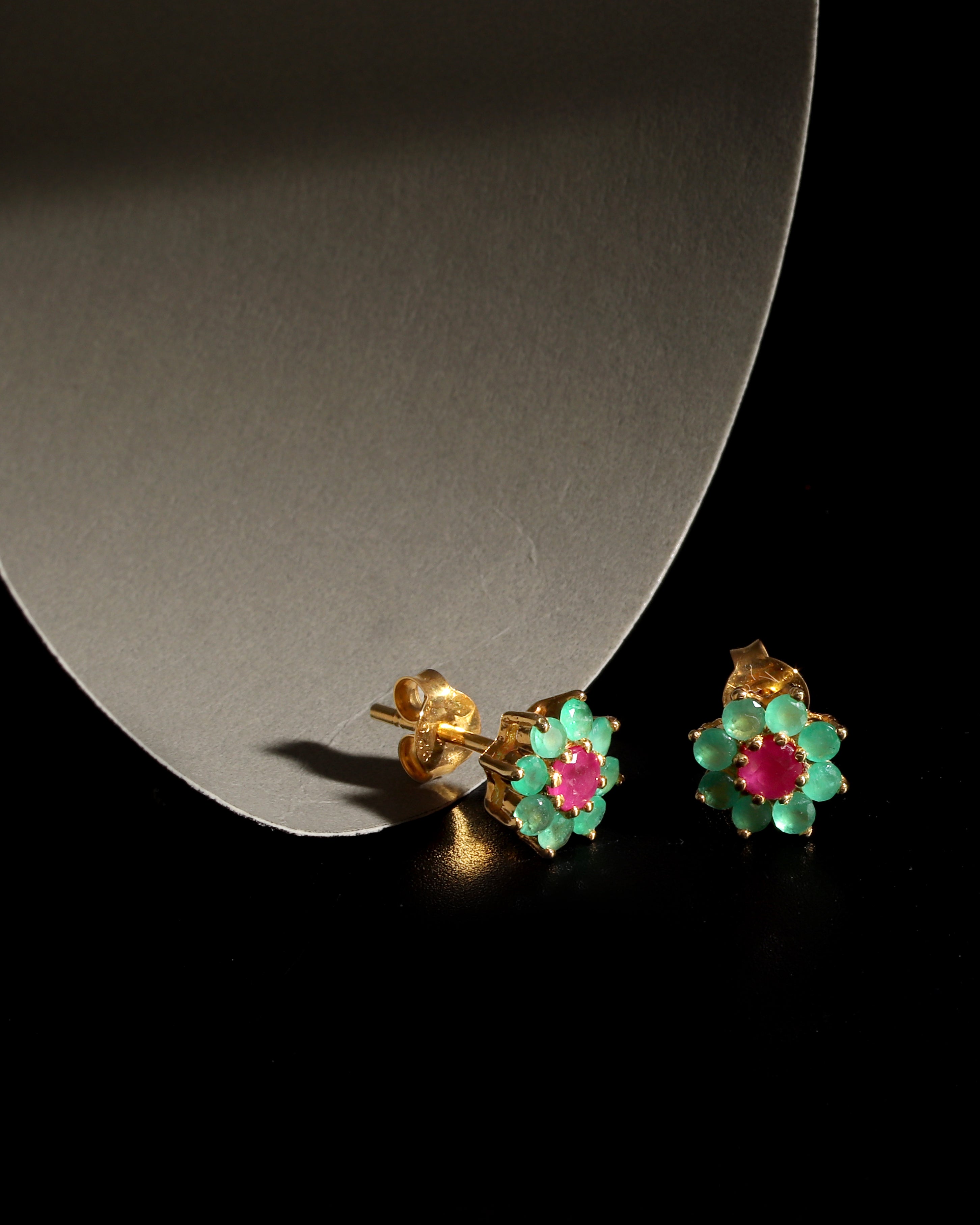 A pair of gold flower-shaped stud earrings with green enamel petals and pink centers, displayed on a black surface next to a curved gray stand.