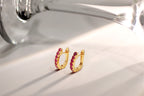 A pair of small gold huggie hoop earrings set with bright pink gemstones, resting on a white surface with soft shadows and a blurred glass in the background.