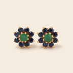 A pair of gold flower-shaped stud earrings featuring a central green oval stone surrounded by six dark blue round stones, set against a plain beige background.