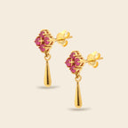 A pair of gold dangle earrings with a four-petal flower design set with pink gemstones, hanging from teardrop-shaped gold drops, shown on a light beige background.
