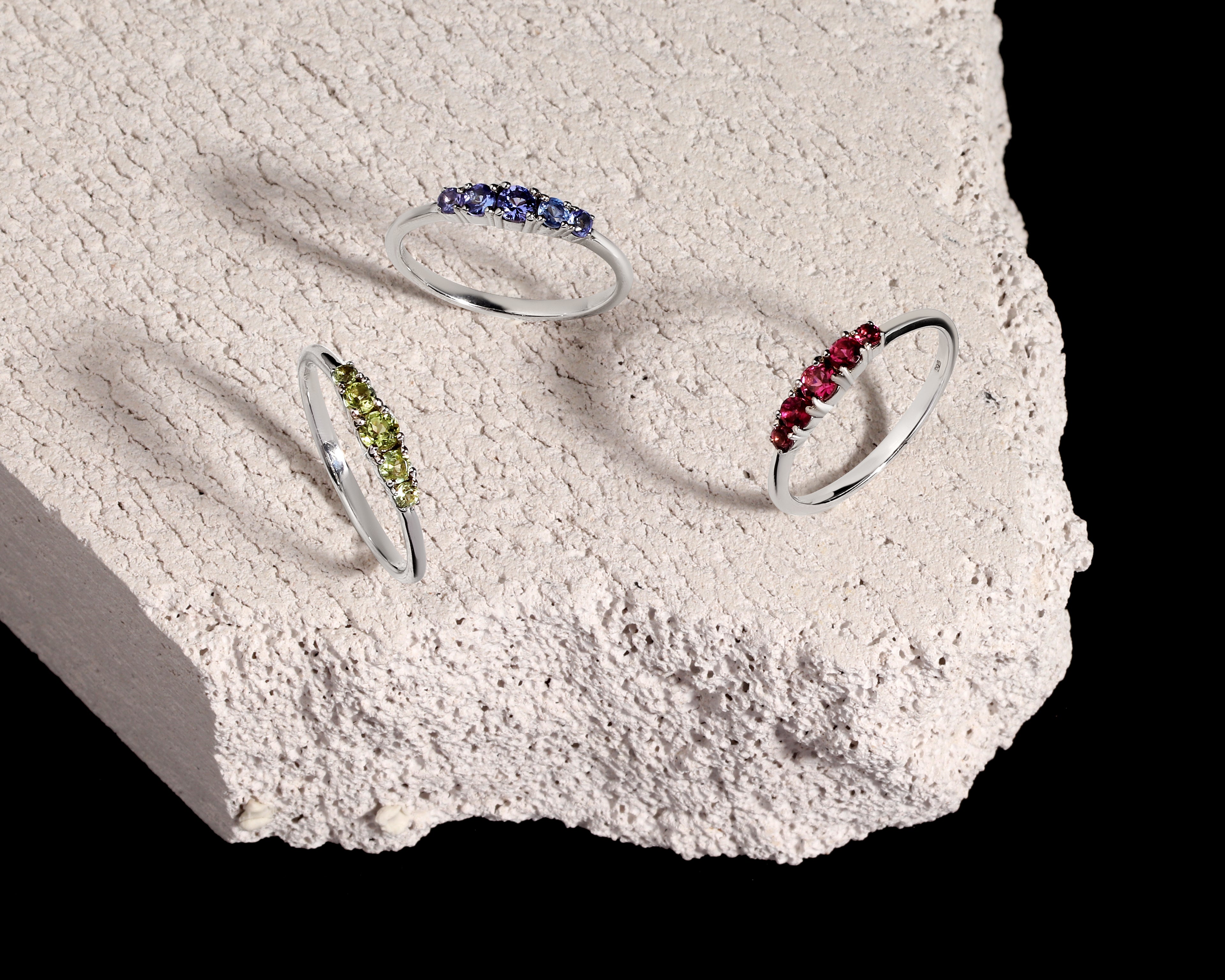 Three silver rings with small colored gemstone clusters are displayed on a rough white stone slab against a black background.