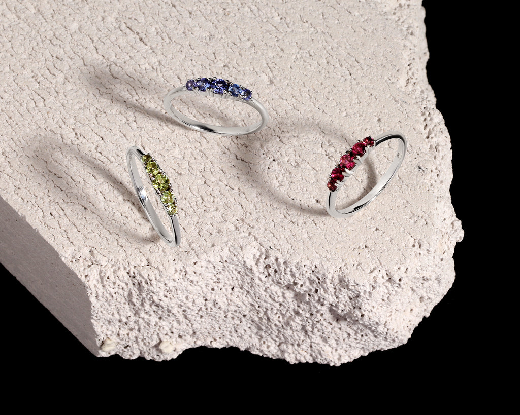 Three silver rings with small colored gemstone clusters are displayed on a rough white stone slab against a black background. 