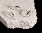 Three silver rings with small colored gemstone clusters are displayed on a rough white stone slab against a black background. 