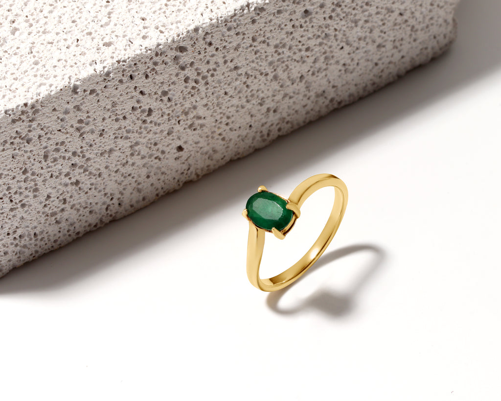 A gold ring with an oval-cut green gemstone, displayed on a clean white surface next to a textured gray stone block.
