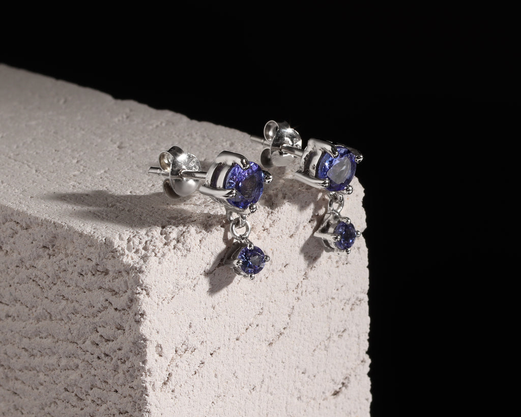 A pair of silver stud earrings with oval blue gemstones and small dangling teardrop-shaped blue gemstones, displayed on a textured white block against a solid black background.