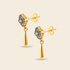 A pair of gold dangle earrings with blue gemstone floral clusters and teardrop-shaped gold drops, shown on a light beige background.