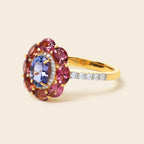 A yellow gold ring featuring a floral cluster setting with a central oval blue sapphire surrounded by round pink garnet stones, with a row of small clear stones along the band, set against a solid light beige background.