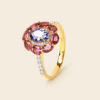 A yellow gold ring featuring a flower-shaped cluster with pink gemstones, a central blue gemstone, and a halo of small clear stones, set against a light beige background.