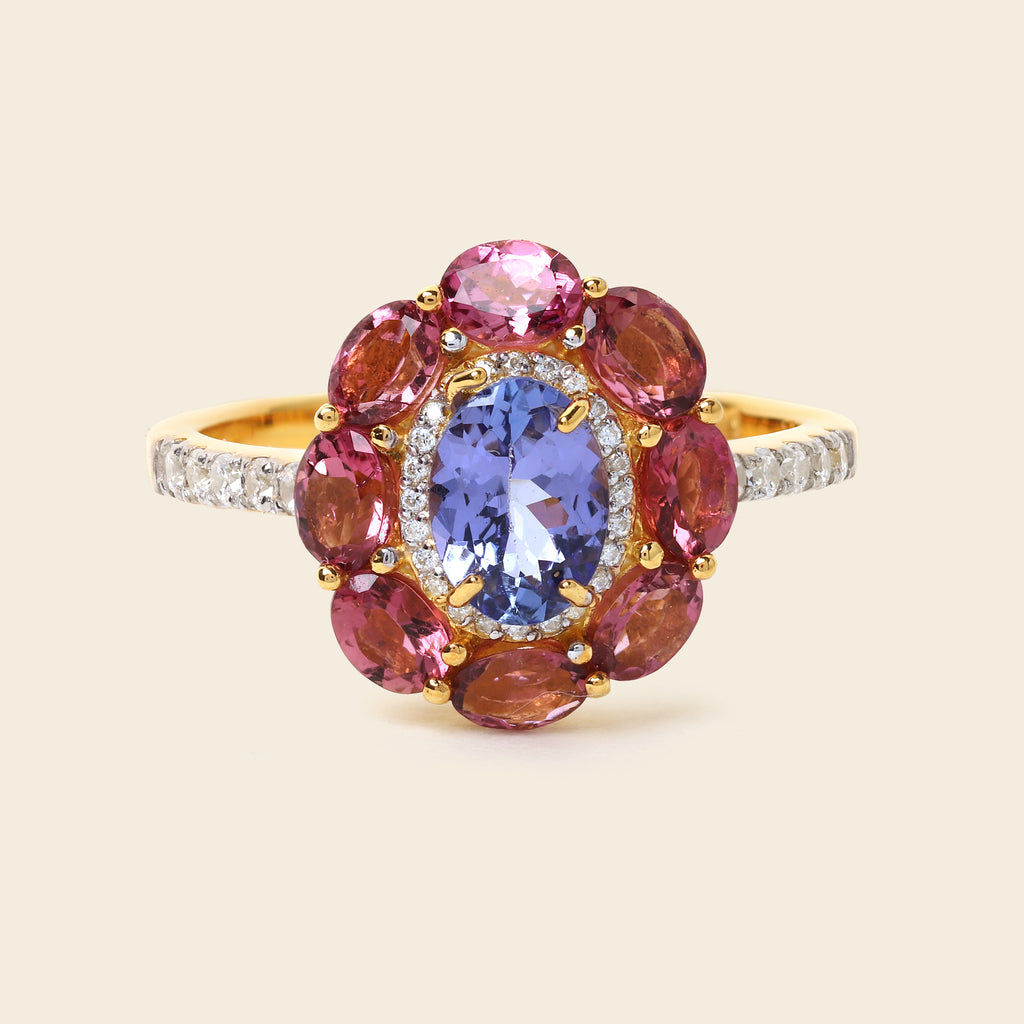 A yellow gold ring featuring a central oval blue tanzanite gemstone surrounded by a halo of round pink and reddish-brown garnet-like stones, with small clear stones along the band, set against a solid light beige background.