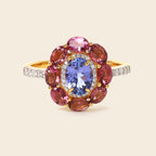 A yellow gold ring featuring a central oval blue tanzanite gemstone surrounded by a halo of round pink and reddish-brown garnet-like stones, with small clear stones along the band, set against a solid light beige background.