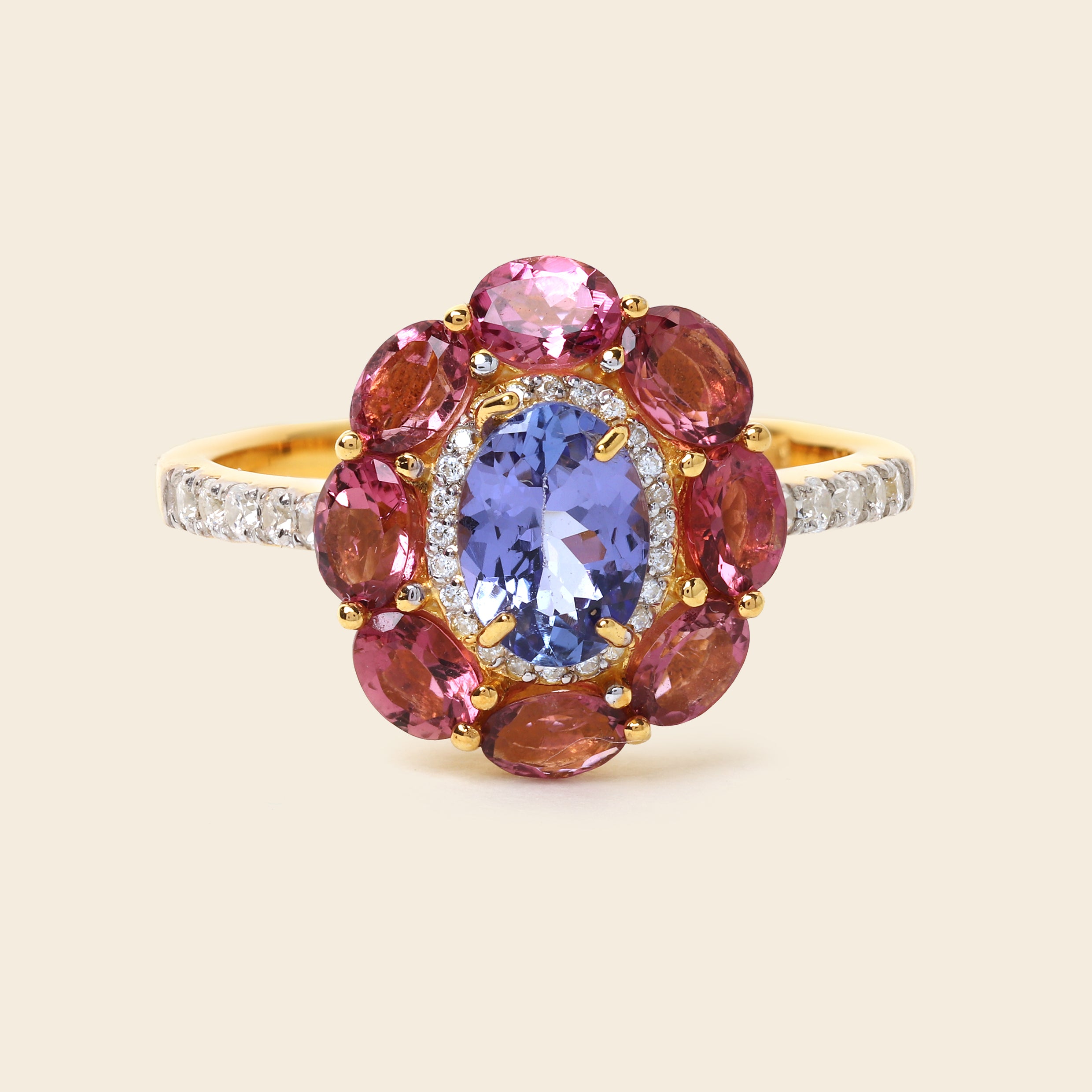A yellow gold ring featuring a central oval blue tanzanite gemstone surrounded by a halo of round pink and reddish-brown garnet-like stones, with small clear stones along the band, set against a solid light beige background.