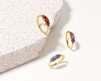 Three gold rings with marquise-cut gemstones in red, purple, and blue, arranged on a white surface next to a textured white stone slab.