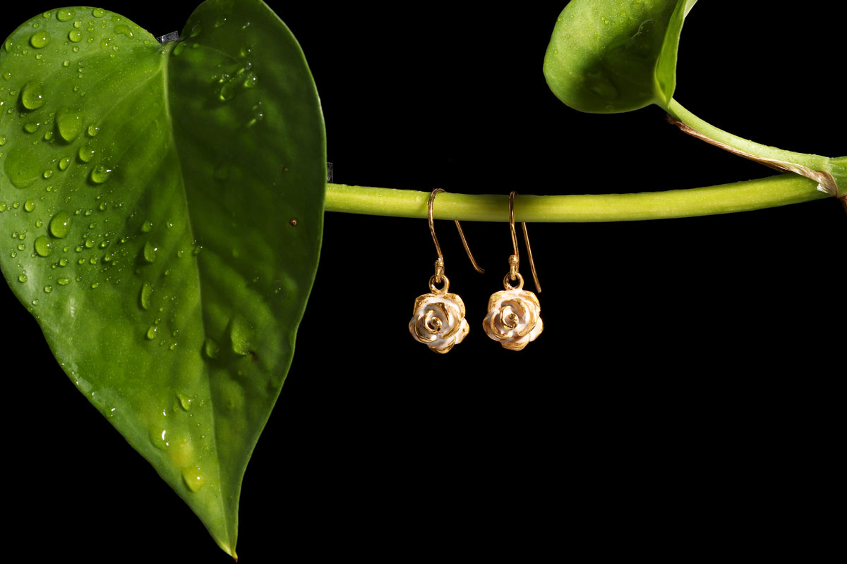 A pair of gold rose-shaped dangle earrings hanging from a green stem against a solid black background.