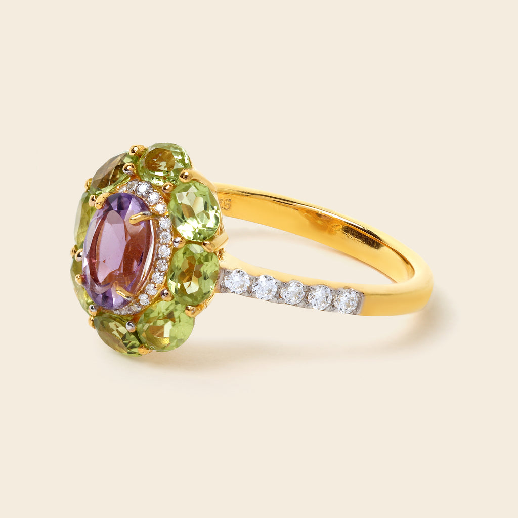 A yellow gold ring featuring a central oval purple amethyst gemstone surrounded by a halo of green peridot gemstones, with a pavé band set with small clear round-cut diamonds, shown on a light beige background.