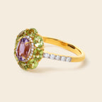 A yellow gold ring featuring a central oval purple amethyst gemstone surrounded by a halo of green peridot gemstones, with a pavé band set with small clear round-cut diamonds, shown on a light beige background.