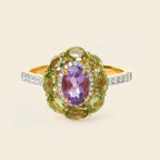 A yellow gold ring featuring a central oval purple amethyst gemstone surrounded by a halo of green peridot gemstones, with a pavé band set with small clear round-cut stones, shown on a light beige background.