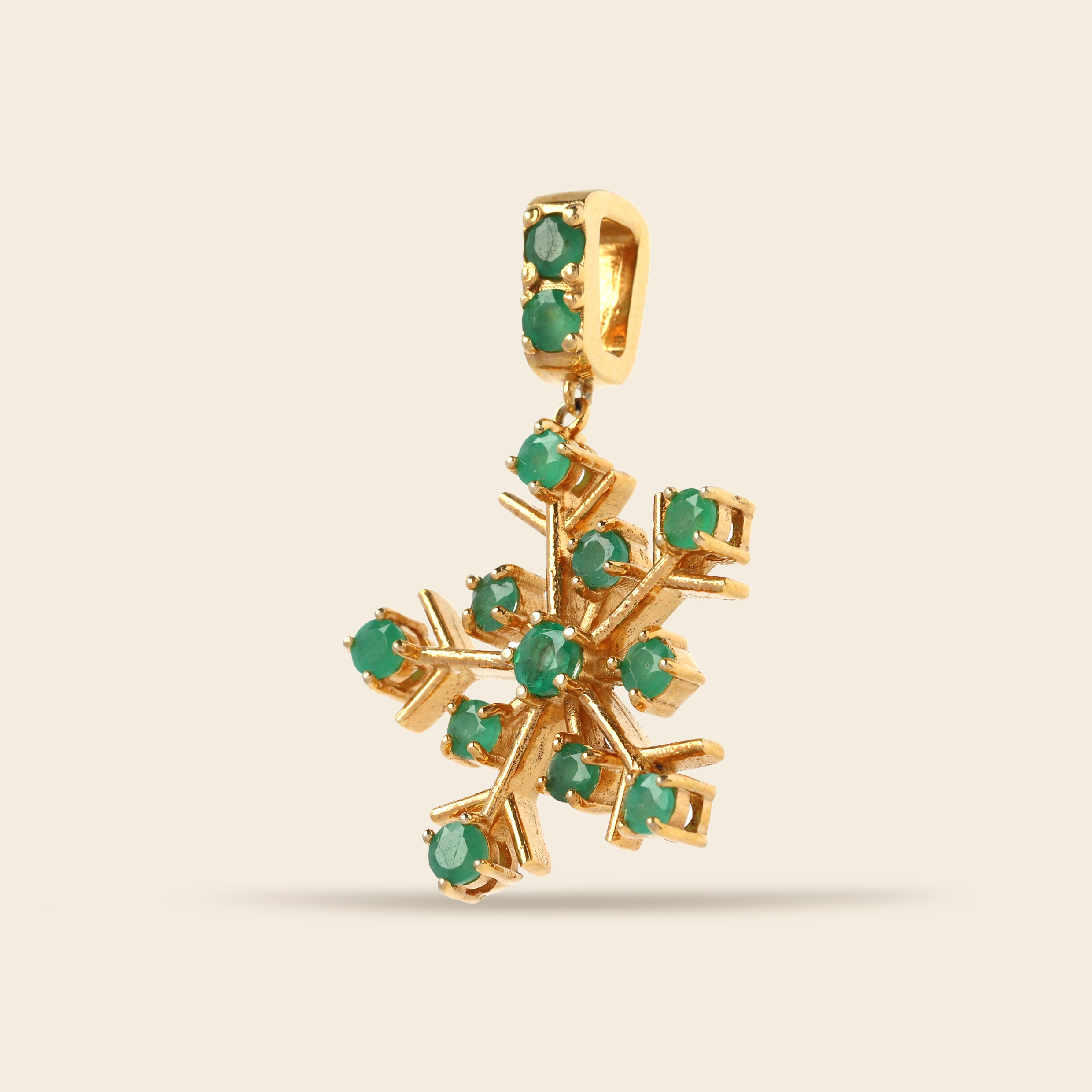 A gold snowflake pendant charm embellished with small round green gemstones, shown against a solid light beige background.