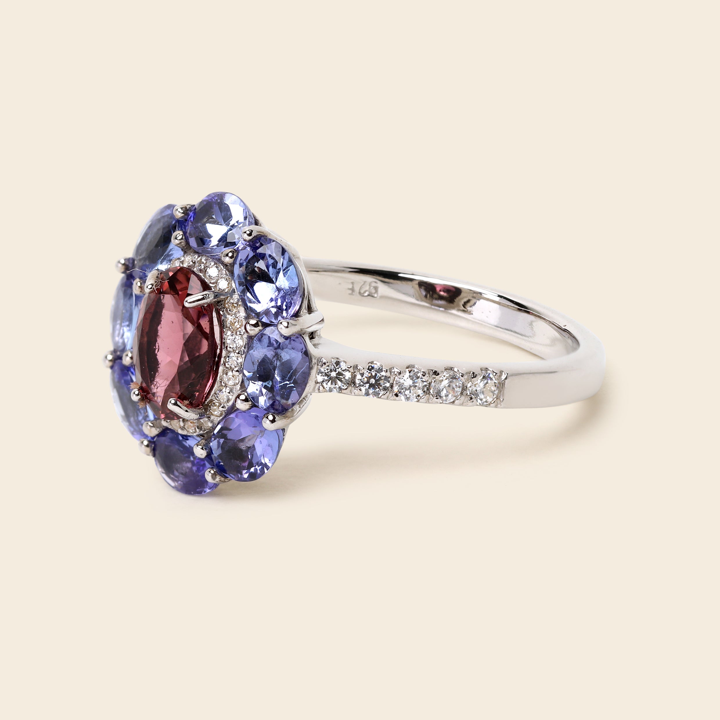 A silver ring featuring a floral cluster setting with a central marquise-cut pinkish-red gemstone surrounded by round blue gemstones, with a pavé band on a light beige background.