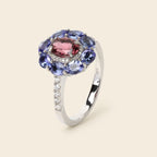 A silver ring featuring a central oval pink gemstone surrounded by a halo of round blue gemstones, with a slim pavé-set silver band, shown on a light beige background.