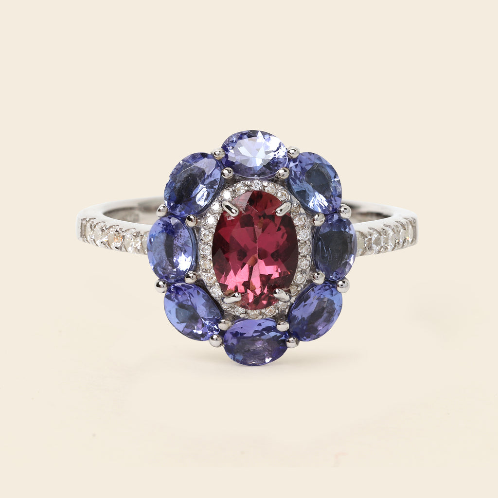 A silver-tone ring featuring a central oval pinkish-red gemstone surrounded by a halo of round blue gemstones and small clear accent stones, shown on a light beige background.