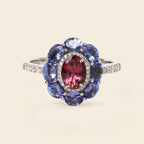 A silver-tone ring featuring a central oval pinkish-red gemstone surrounded by a halo of round blue gemstones and small clear accent stones, shown on a light beige background.