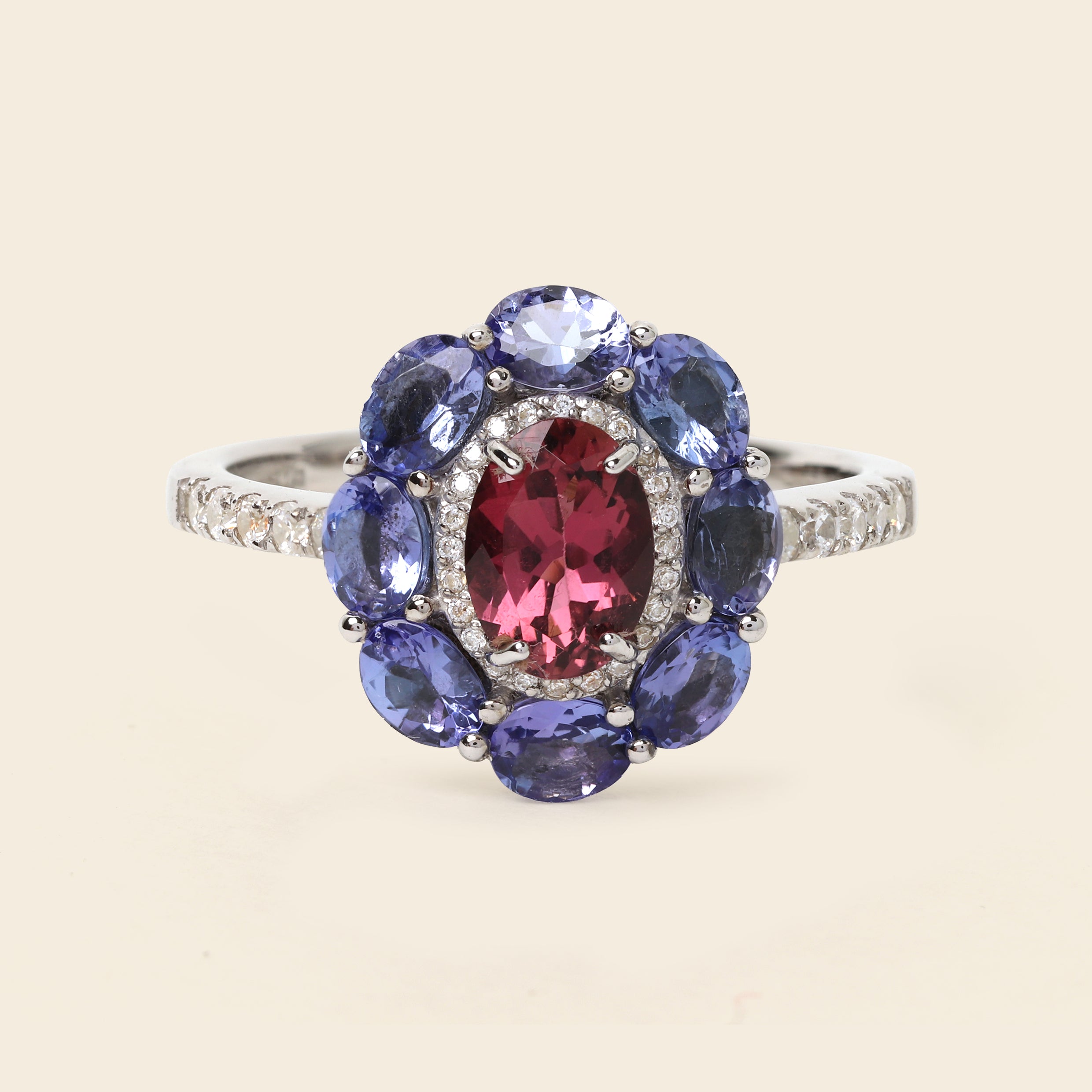 A silver-tone ring featuring a central oval pinkish-red gemstone surrounded by a halo of round blue gemstones and small clear accent stones, shown on a light beige background.