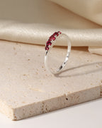 A silver ring with a curved row of small red gemstones, displayed on a textured beige stone slab with cream-colored fabric in the background.