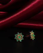 A pair of gold-toned floral stud earrings with green gemstone centers and surrounding stones, displayed on a black surface with a draped dark red satin fabric background.