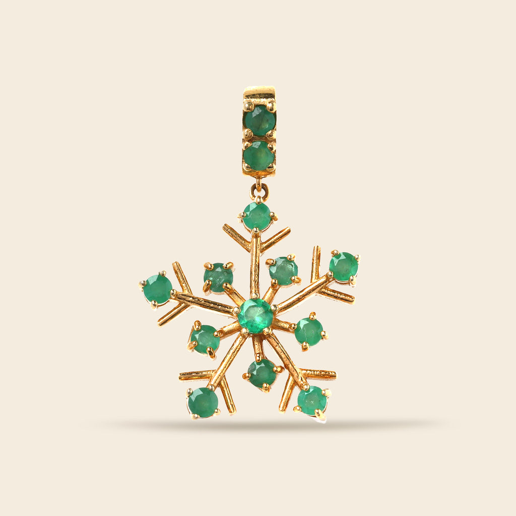 A gold snowflake-shaped pendant featuring round green gemstones set along its points and in the bail, shown against a solid light beige background.