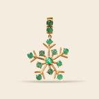 A gold snowflake-shaped pendant featuring round green gemstones set along its points and in the bail, shown against a solid light beige background.