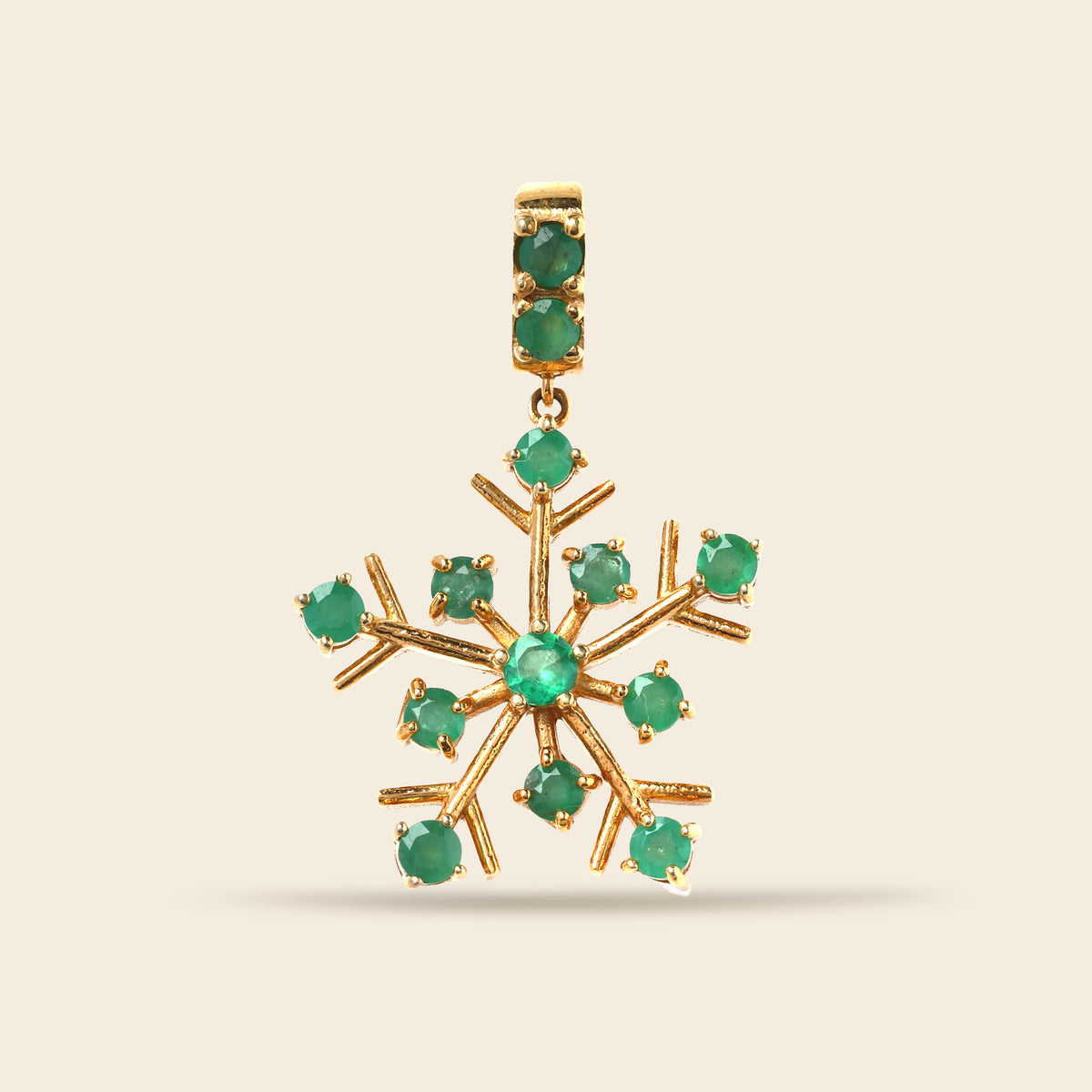 A gold snowflake-shaped pendant featuring round green gemstones set along its points and in the bail, shown against a solid light beige background.