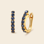 A pair of gold huggie hoop earrings with a row of round-cut dark blue sapphires, shown on a light beige background.