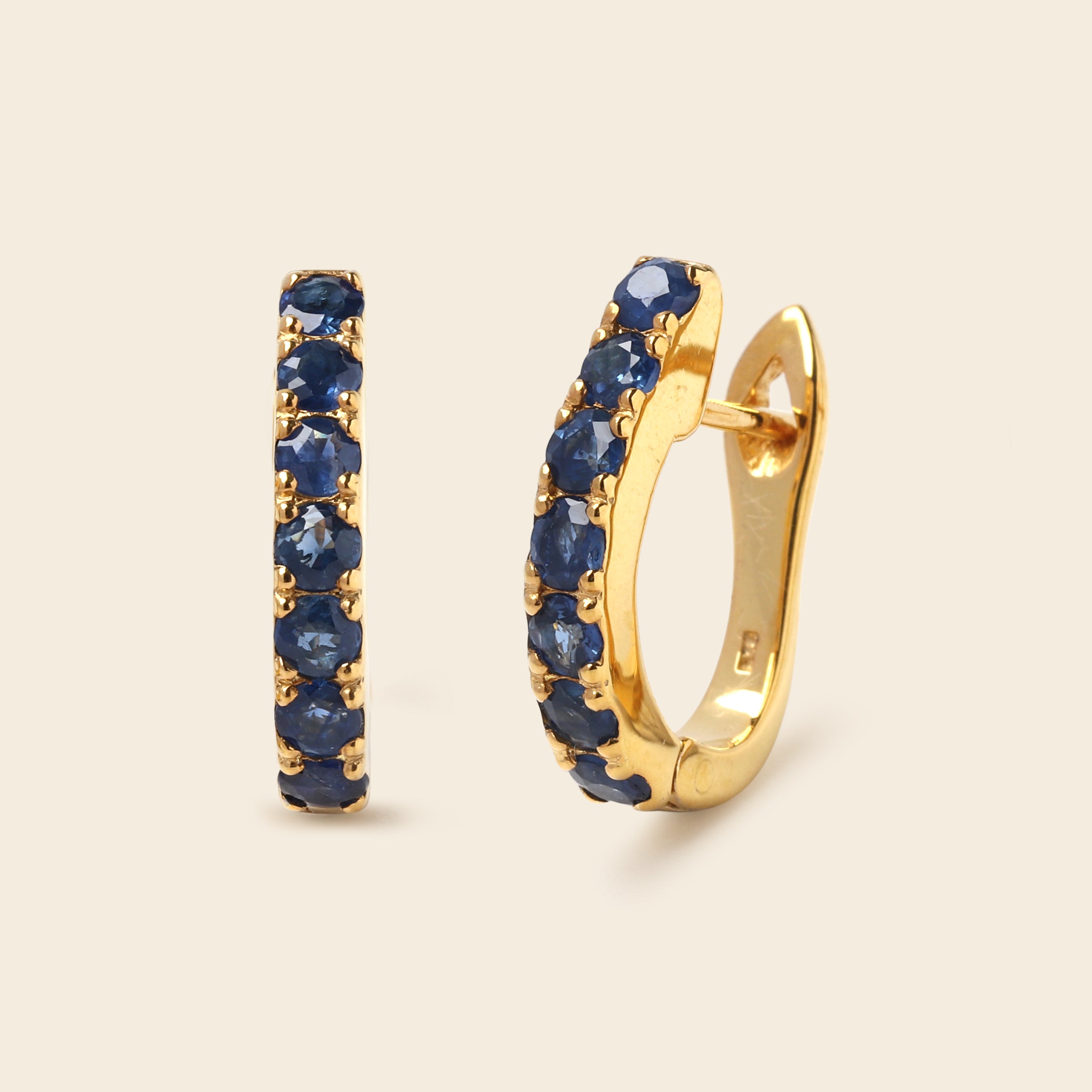 A pair of gold huggie hoop earrings with a row of round-cut dark blue sapphires, shown on a light beige background.