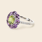 A silver ring featuring a central oval green peridot gemstone surrounded by a halo of round purple amethysts and small clear accent stones on the band, set against a solid light beige background.