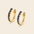 A pair of gold huggie hoop earrings embellished with a row of deep blue round gemstones, set against a solid light beige background.