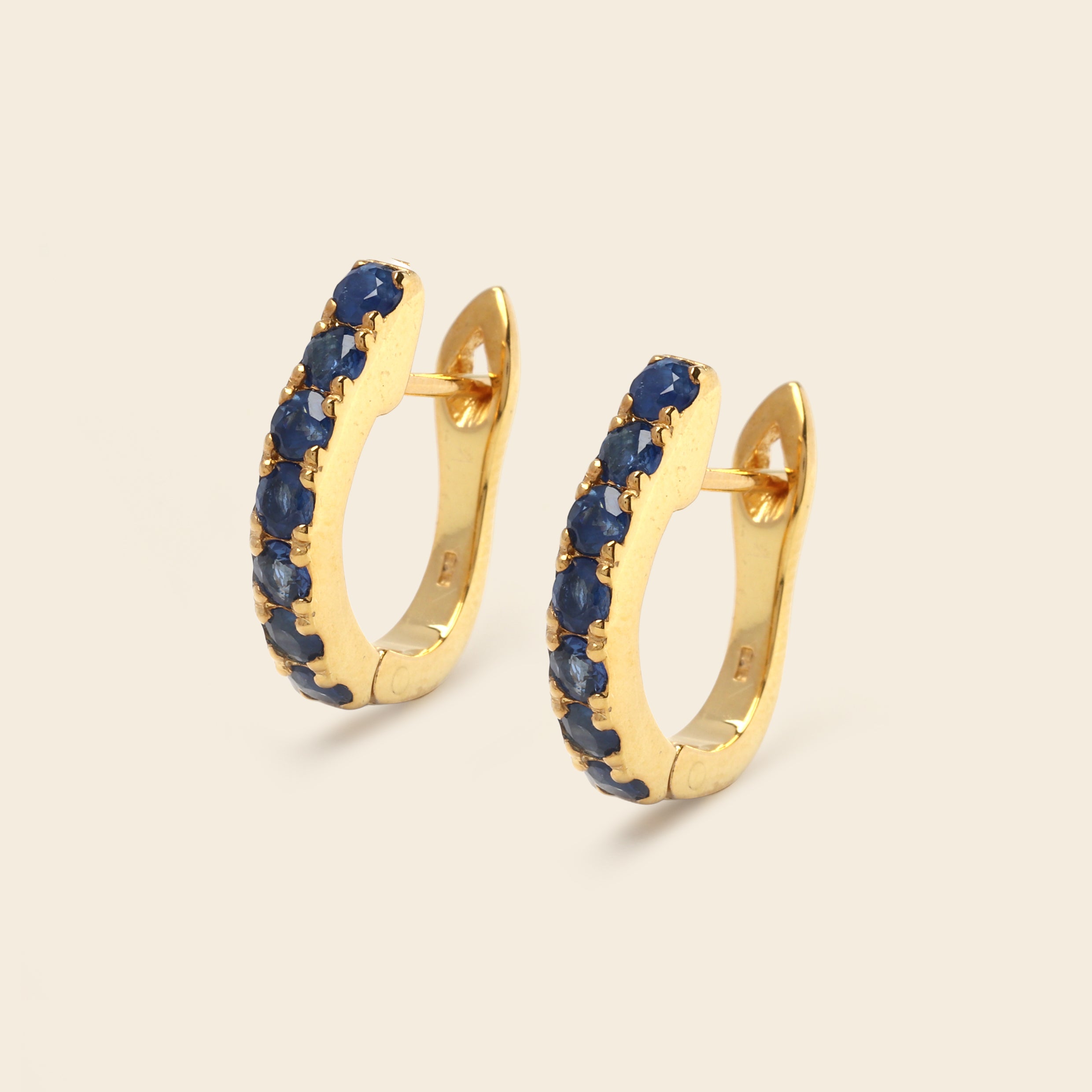 A pair of gold huggie hoop earrings embellished with a row of deep blue round gemstones, set against a solid light beige background.