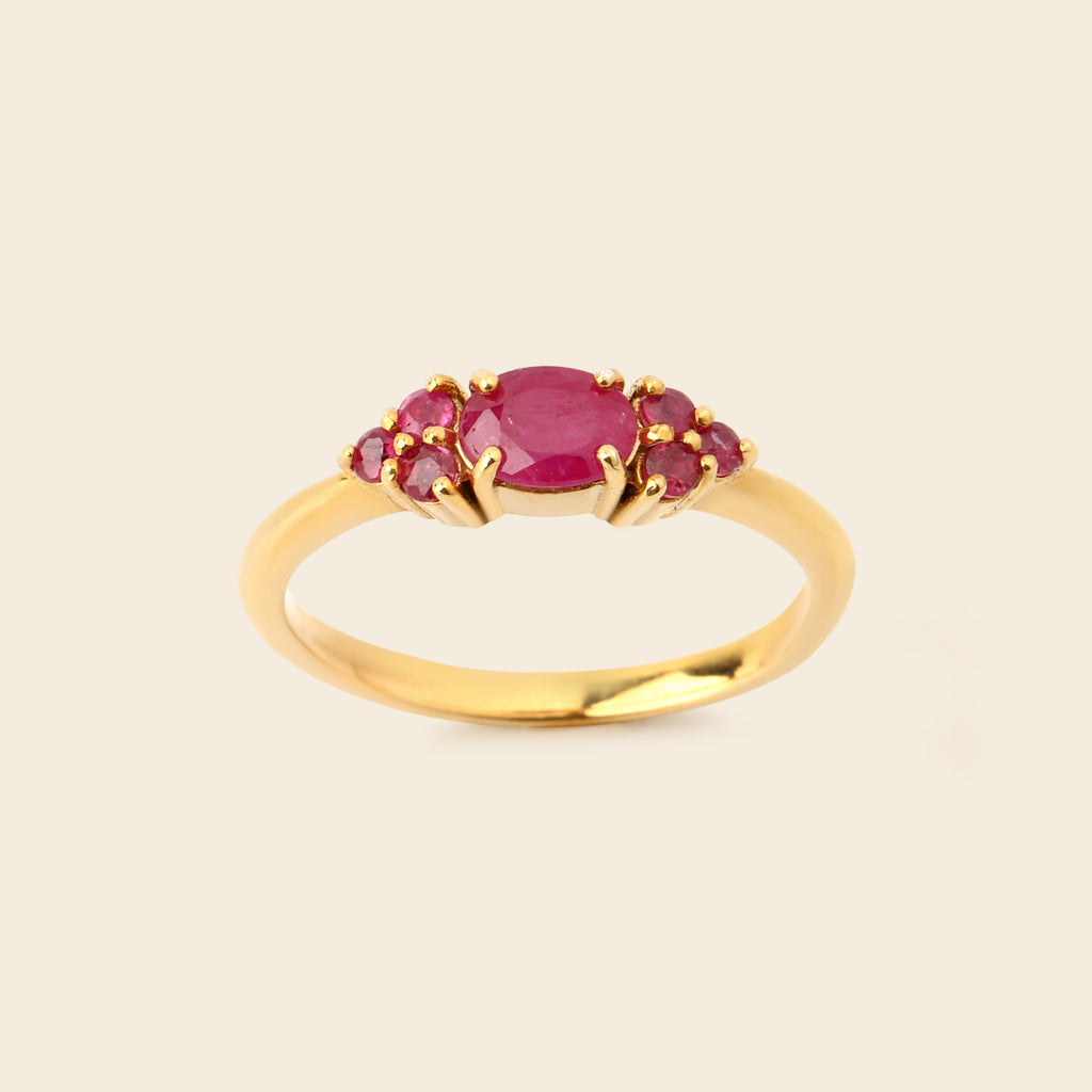 A yellow gold ring featuring an oval-cut pinkish-red center stone flanked by three smaller round stones, set against a solid light beige background.
