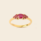 A yellow gold ring featuring an oval-cut pinkish-red center stone flanked by three smaller round stones, set against a solid light beige background.