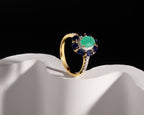 A gold ring with a round green center stone and a halo of deep blue and small clear stones, displayed on a white sculptural surface against a black background.