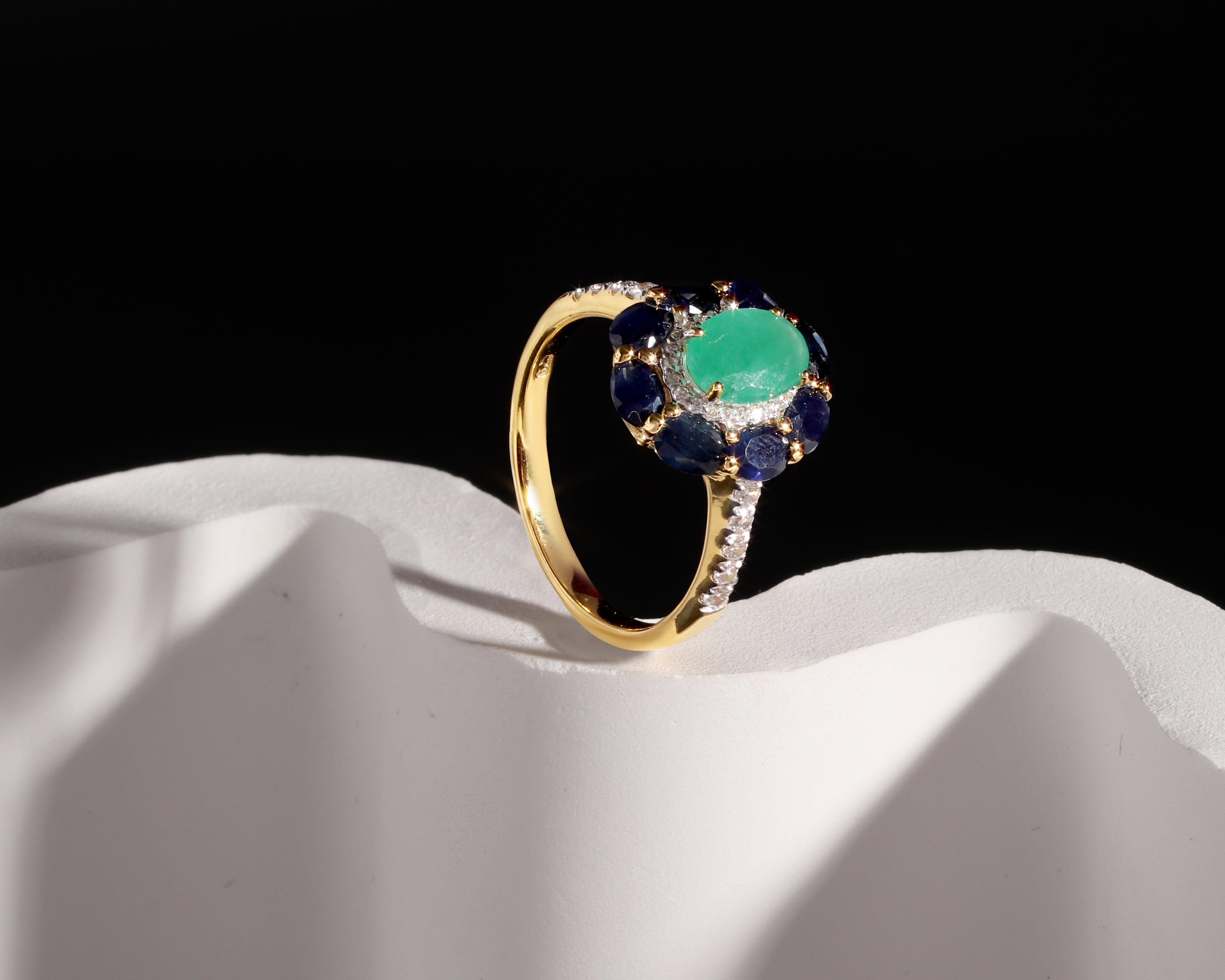 A gold ring with a round green center stone and a halo of deep blue and small clear stones, displayed on a white sculptural surface against a black background.