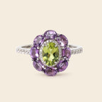 A floral cluster ring featuring a central oval green peridot gemstone surrounded by a halo of round purple amethyst stones, set in a silver-colored band with small pavé diamonds, on a solid light beige background.