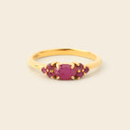 A gold ring featuring a central oval pinkish-red gemstone flanked by smaller round pinkish-red stones, set against a plain light beige background.