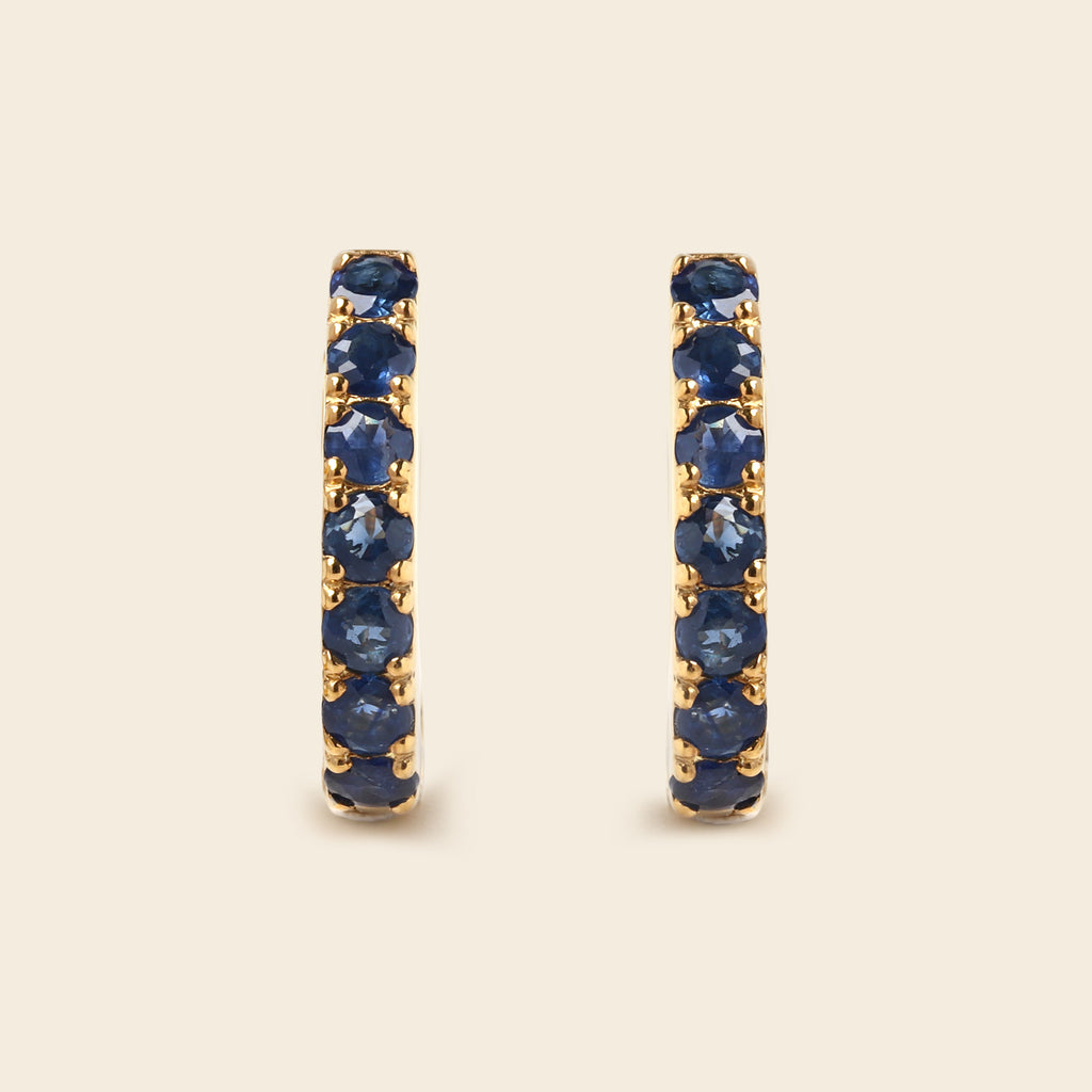 A pair of gold huggie hoop earrings set with round-cut deep blue sapphires, displayed on a light beige background.