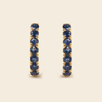A pair of gold huggie hoop earrings set with round-cut deep blue sapphires, displayed on a light beige background.