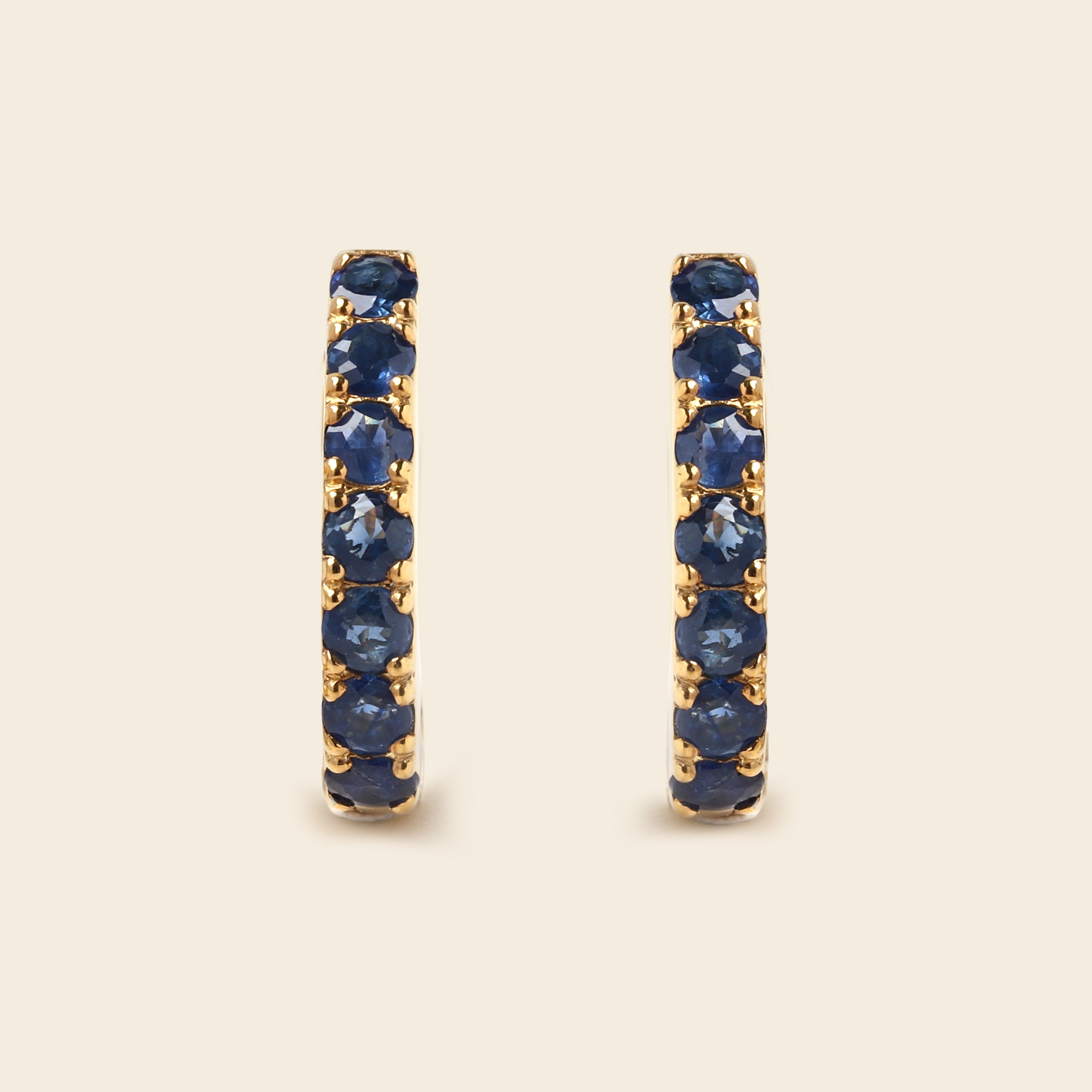 A pair of gold huggie hoop earrings set with round-cut deep blue sapphires, displayed on a light beige background.