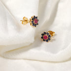 A pair of gold flower-shaped stud earrings with dark blue gemstones and a central pink gemstone, displayed on a soft white fabric background.