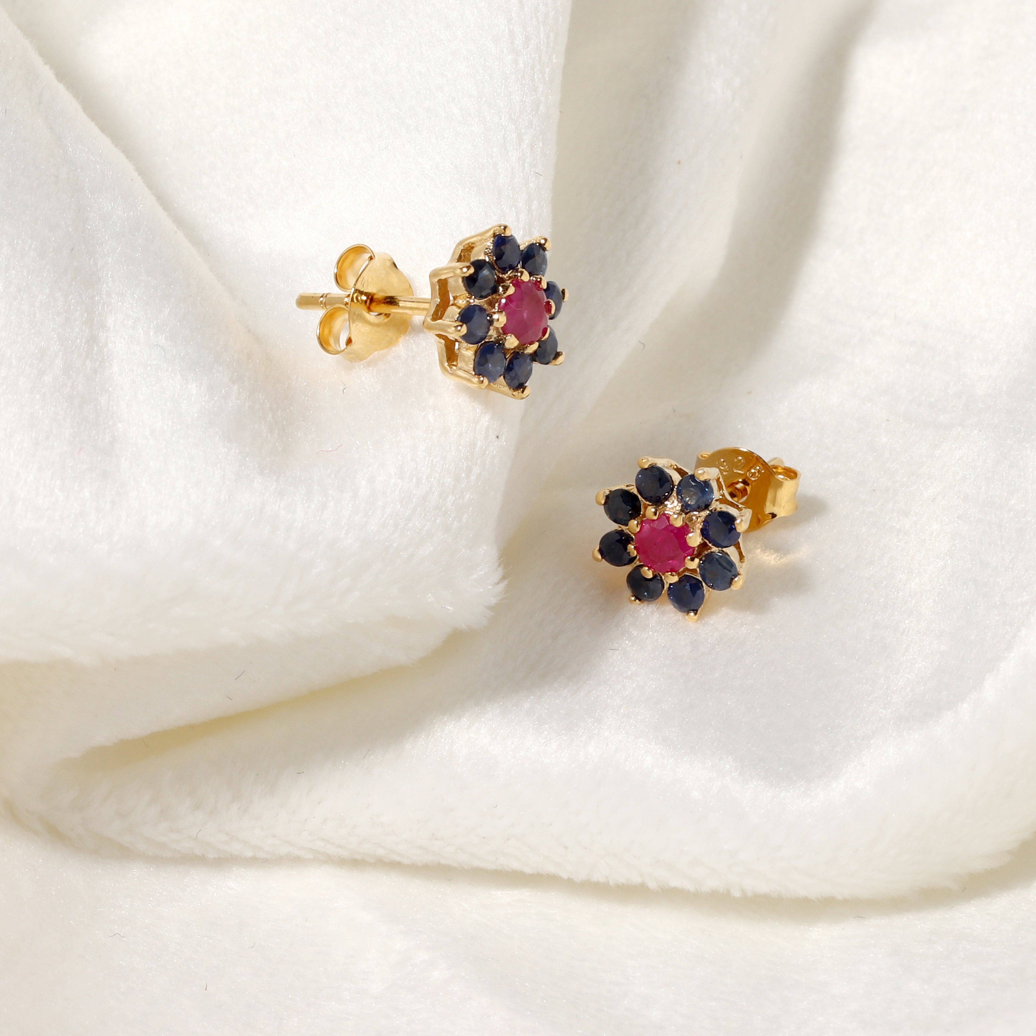 A pair of gold flower-shaped stud earrings with dark blue gemstones and a central pink gemstone, displayed on a soft white fabric background.