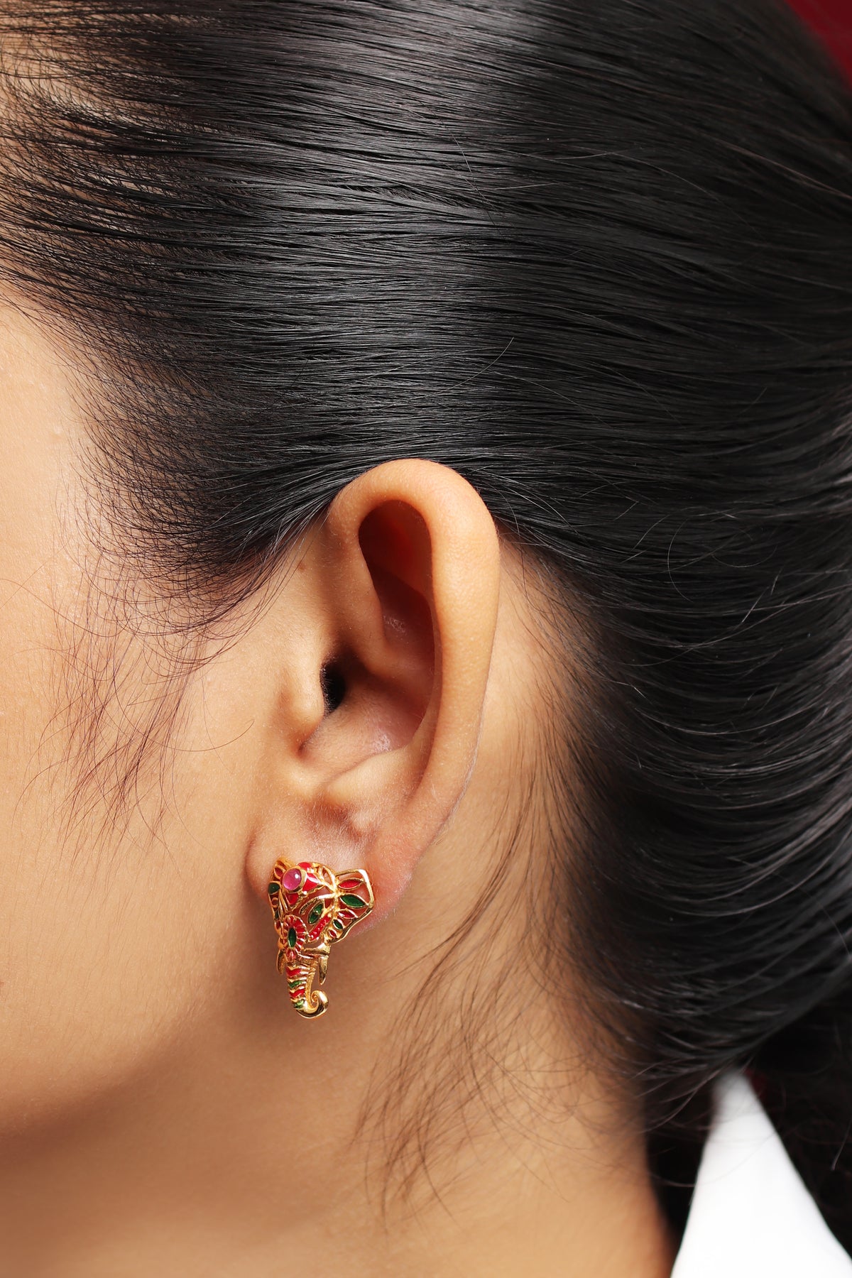 A close-up of a person's ear wearing a small elephant-shaped stud earring with red and green enamel details and gold-toned metal.