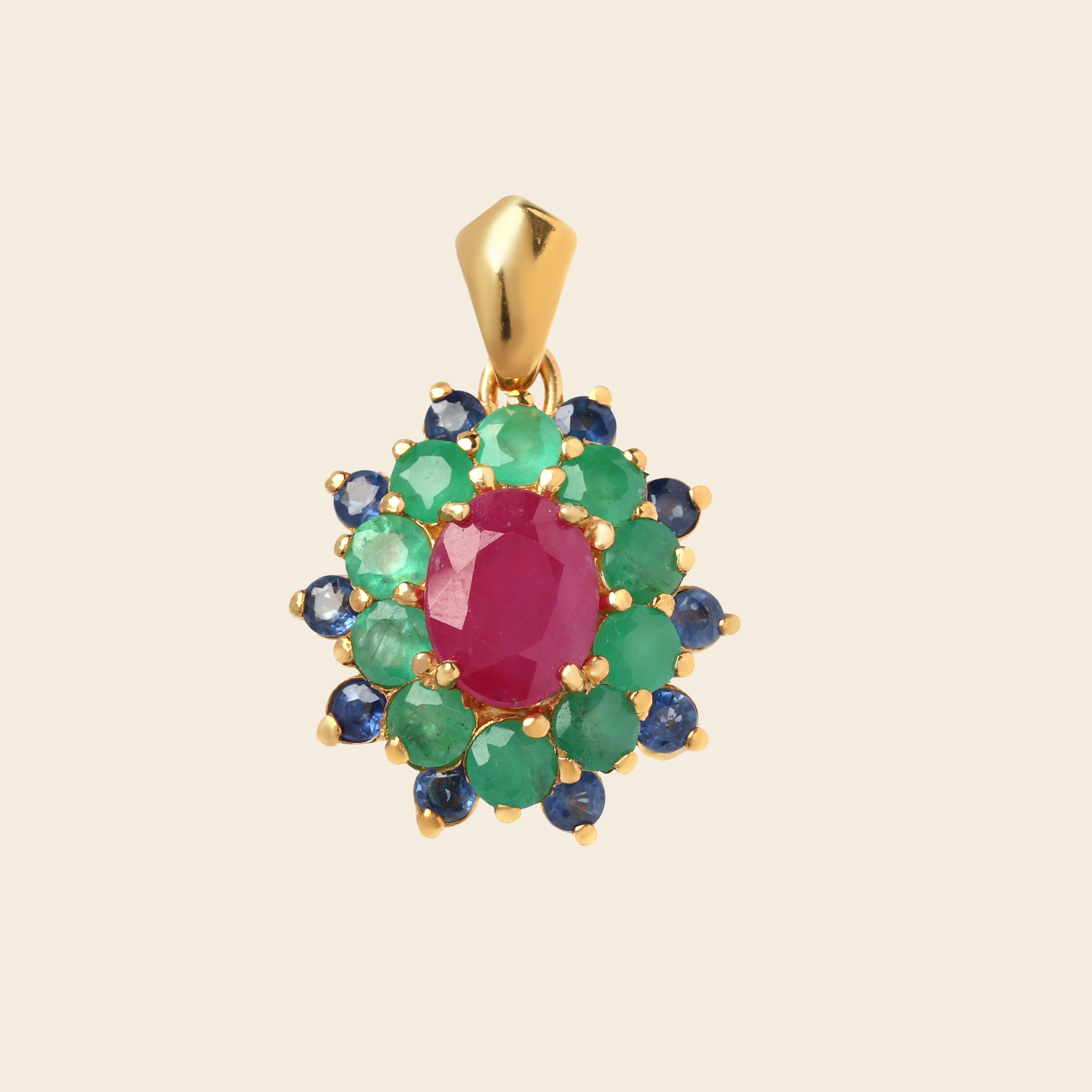 A gold pendant featuring a central oval pinkish-red ruby gemstone surrounded by a double halo of round green emeralds and blue sapphires, set in yellow gold on a light beige background.