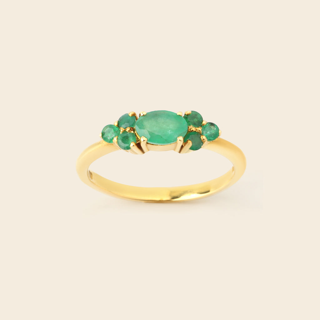 A gold ring featuring a central oval green emerald-cut stone surrounded by four smaller round green gemstones, set on a smooth gold band against a light beige background.